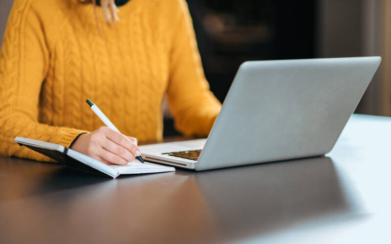 Woman writing in a notebook with a laptop open in front of her